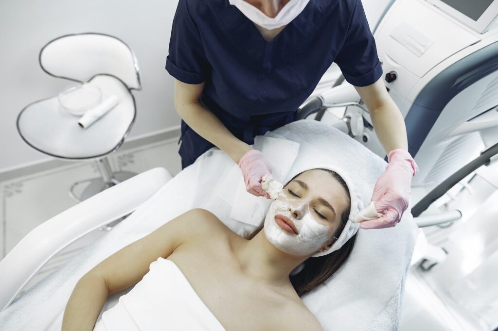 crop female cosmetologist removing purifying mask from face of relaxed woman in contemporary clinic